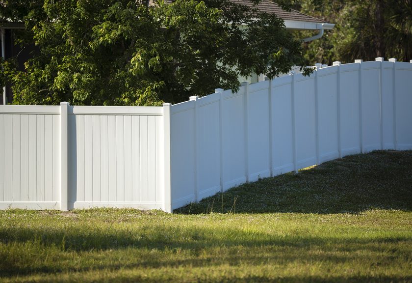 White vinyl privacy fence running along a sloped lawn under shade trees
